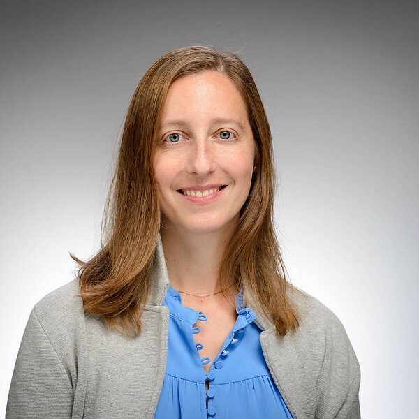 Woman with brown hair and blue eyes smiles, wearing a blue shirt and gray cardigan.